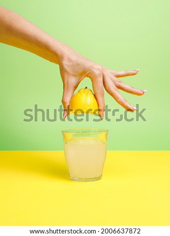 A woman's hand squeezing half of a lemon and making a glass of icy cold lemonade isolated on a yellow and mint green background. Cocktail ingredient. Creative summer refreshing drink concept Royalty-Free Stock Photo #2006637872