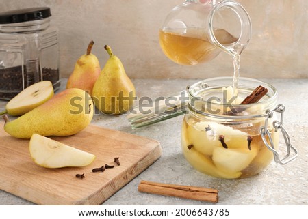 Pouring flavored sugar water in a glass jar with sliced pear, cinnamon and cloves on rustic stone, canning summer fruits for the winter time, copy space, selected focus, narrow depth of field 