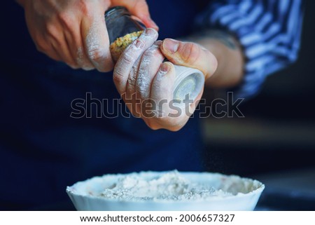 A chef in a blue apron and striped shirt holds a glass salt shaker with seasoning and pours it into a bowl of flour for future baking. Home cooking.