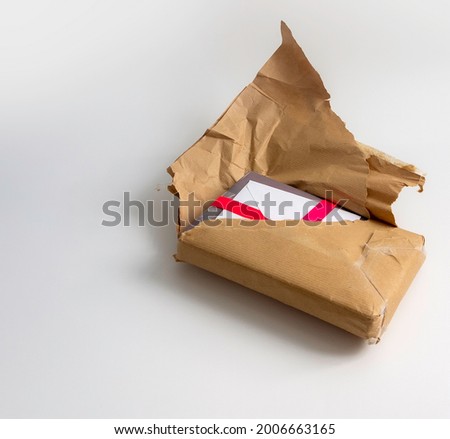 Torn brown postal parcel package revealing wrapped gift or present that is with a pink satin ribbon and a card inside a white envelope. Text space, isolated on white background. Opening gift concept.