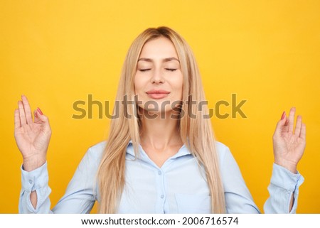 Businesswoman in a blue shirt keeps eyes closed holding fingers in mudra gesture, beautiful manager girl keeps calm relaxes isolated on yellow background, anti-stress therapy meditation