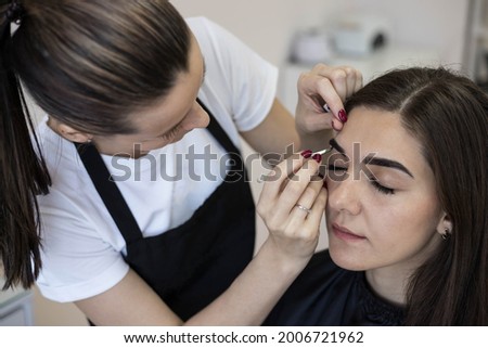 The master brow artist plucks the eyebrows with tweezers to the client. The master corrects the shape of the eyebrows with tweezers after waxing Royalty-Free Stock Photo #2006721962