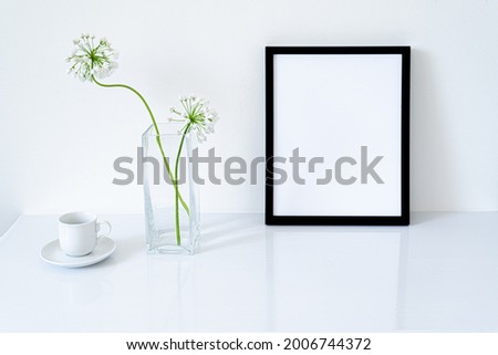 Blank black frame mockup. Small fresh flowers of white garlic (allium neapolitanum) in glass vase on glossy white table. White background, minimal and elegant space
