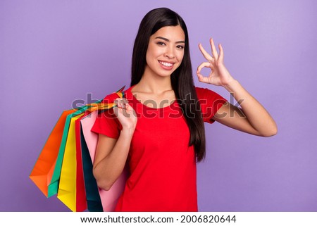 Photo of adorable pretty young lady wear red outfit smiling showing okey holding back shopping bags isolated violet color background