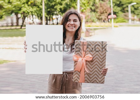 Young woman holding a white poster in her hands and packaging with a bow and smiling against the background of the park