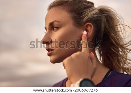 Serious young sporty female touching TWS earbuds while listening to music during outdoor fitness training Royalty-Free Stock Photo #2006969276