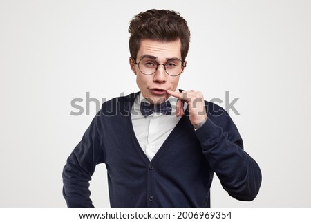 Funny young male student in elegant suit and nerdy glasses touching lips and looking at camera against white background
