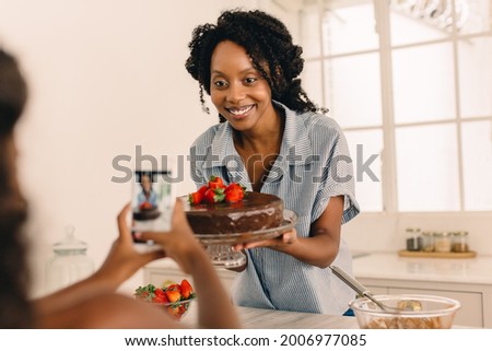Woman holding a cake being photographed by her daughter at home. Girl taking photo with mobile phone of her mother with cake in kitchen.
