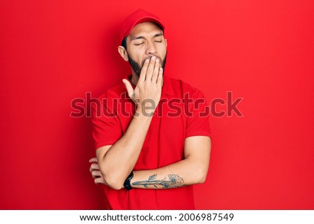 Hispanic man with beard wearing delivery uniform and cap bored yawning tired covering mouth with hand. restless and sleepiness. 