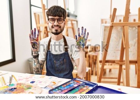 Hispanic man with beard at art studio showing and pointing up with fingers number nine while smiling confident and happy. 