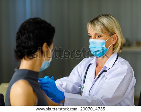 Beautiful short black hair female patient wears face mask sit look at camera while Caucasian doctor in white lab coat with stethoscope injecting vaccine in to her shoulder in blurred background.