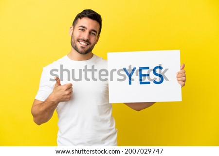 Young handsome caucasian man isolated on yellow background holding a placard with text YES with thumb up