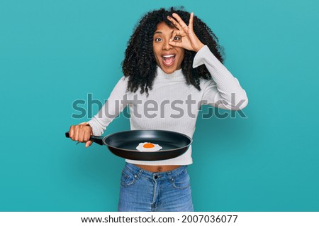 Young african american girl holding skillet with fried egg smiling happy doing ok sign with hand on eye looking through fingers 