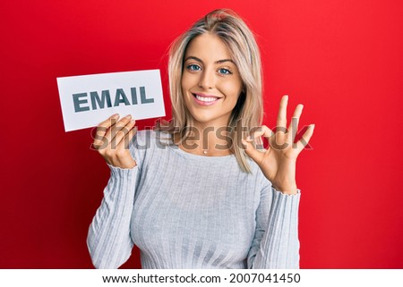 Beautiful blonde woman holding paper with email address doing ok sign with fingers, smiling friendly gesturing excellent symbol 
