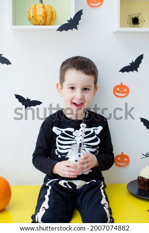 Toddler caucasian boy in skeleton costume sits on the table with Halloween food and decorations and drinks milk from bottle with eyes