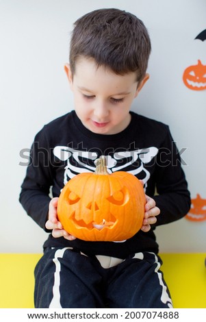Toddler caucasian boy in skeleton costume holds curved pumpkin and sits on the table with Halloween food and decorations