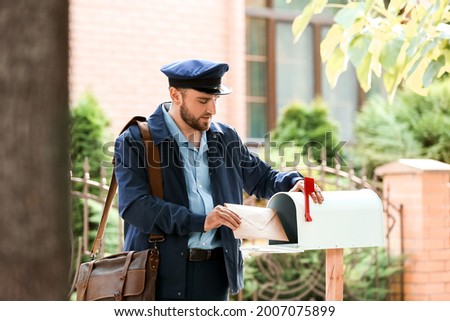 Handsome young postman putting letter in mail box outdoors Royalty-Free Stock Photo #2007075899