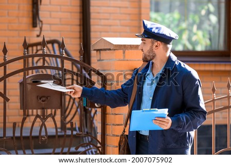 Handsome young postman putting letter in mail box outdoors Royalty-Free Stock Photo #2007075902