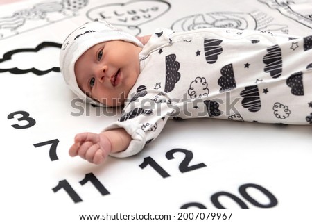 Full length portrait of baby wearing  slips and a hat with a black and white pattern lying on a rug with a black and white picture of months from birth