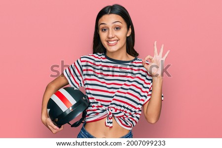 Young hispanic girl holding motorcycle helmet doing ok sign with fingers, smiling friendly gesturing excellent symbol 
