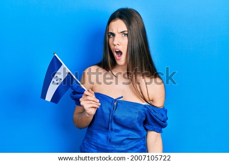 Young brunette teenager holding honduras flag scared and amazed with open mouth for surprise, disbelief face 