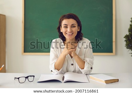 Happy positive school teacher against classroom interior background with blackboard. Beautiful Caucasian woman looking at camera while sitting at desk in online class. Video conference webcam portrait
