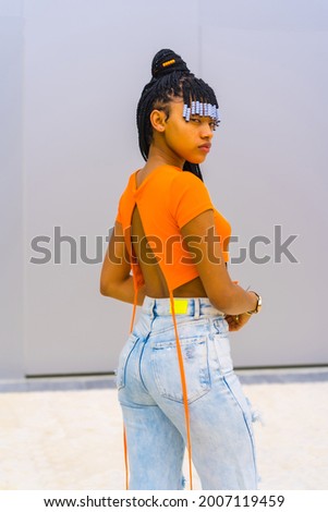 A young trap dancer with braids. Black ethnic girl with orange t-shirt and cowboy pants. Posé with his back
