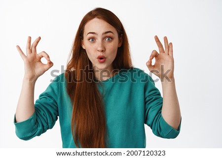 Portrait of impressed redhead girl look in awe, amazed by something cool, show okay OK gesture and recommend, leave positive good feedback, white background
