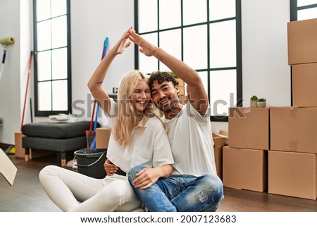 Young beautiful couple doing house symbol with arms raised at new home.