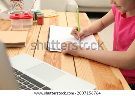 Little girl drawing on paper with pencil at online lesson indoors, closeup. Distance learning