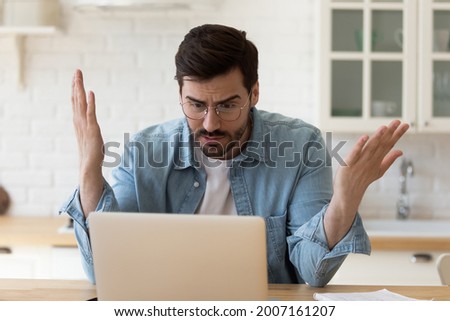 Irritated man in glasses sit at table staring at laptop screen spread arms feels angry and confused having problem with broken computer, reading bad news in email, lost information, app malfunction Royalty-Free Stock Photo #2007161207