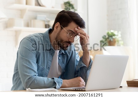 Bad news, lack of understanding of new app, info overload, device malware concept. Young man in glasses sit at table looks at laptop screen touch forehead feels confused has business or pc problems Royalty-Free Stock Photo #2007161291