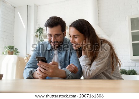 Mixed race couple sit in kitchen at table looking at smartphone screen interested with good e-commerce offer, sale low price and discount in retail services, buying ticket. New mobile app, fun concept