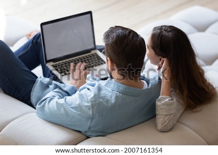 Young spouses sit on sofa relax together at home using computer, buying through e-commerce retail e services, watch new movie online, websurfing, white mock up laptop screen view over couple rear view