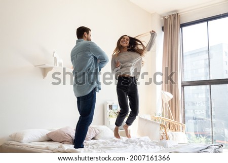 Funny couple jumping on bed in modern bedroom. Cheery happy woman and man in love celebrate relocation day, dance at cozy fashionable home. Tenancy, cohabitation, bank loan for young family concept Royalty-Free Stock Photo #2007161366