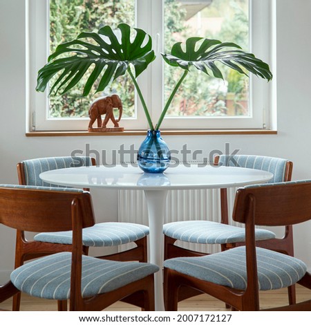 Stylish composition of dining room interior with design table, chairs, tropical leaf in vase, window and elegant decoration in home decor.  Template.
