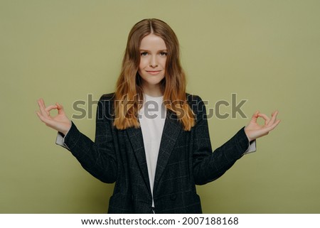 Young beautiful female student wearing in stylish plaid blazer keeping hands in mudra gesture and meditating, feeling calm and happy while practicing meditation, looking at the camera