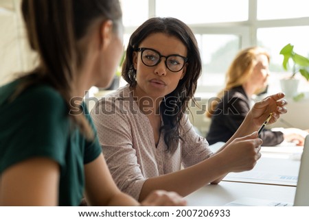 Multiethnic business coworkers discussing startup project. Mixed race mentor training young student, intern, explaining task. Two diverse team members working together, talking at workplace Royalty-Free Stock Photo #2007206933