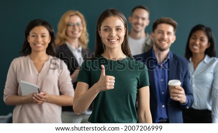 Happy young female business leader making like thumb up, diverse team smiling in background. Satisfied customer, employee with multiethnic group behind giving positive feedback. Head shot portrait