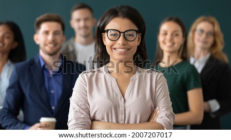Happy mixed raced Black female business leader, confident business woman standing in front of team, smiling at camera. Office employee posing with coworkers in background. Head shot portrait Royalty-Free Stock Photo #2007207017