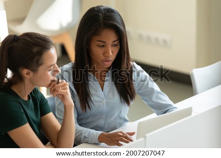 Serious diverse multiethnic student girls, interns collaborating on task at laptop together. Corporate mentor supervising new hired employee, helping with work task. Coworkers discussing project Royalty-Free Stock Photo #2007207047