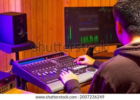 back of asian professional sound engineer mixing audio signal on control surface mixing console in broadcasting, recording studio