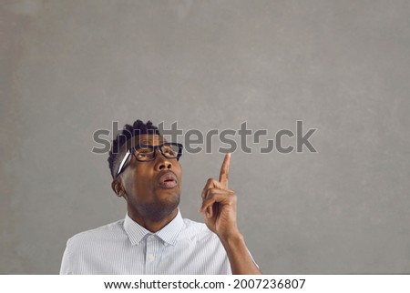 Handsome young black man in white shirt and stylish glasses looks up, points his finger up and gives a whistle of surprise impressed by something on copy space background above