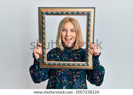 Young blonde woman holding empty frame smiling and laughing hard out loud because funny crazy joke. 