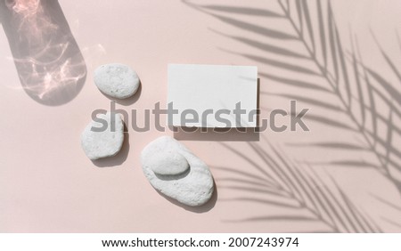 Summer tropical still life mockup composition. Blank invitation card with sea pebbles on beige background. Harsh palm shadows, reflection.  Mockup empty greeting  blank for wedding or business. 