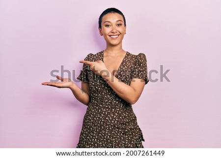 Beautiful hispanic woman with short hair wearing summer dress amazed and smiling to the camera while presenting with hand and pointing with finger. 