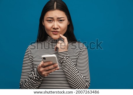 Portrait of a frowning pensive casual young asian woman standing over blue wall background holding mobile phone