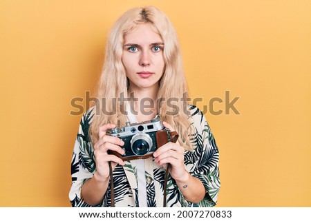 Beautiful caucasian woman with blond hair holding vintage camera relaxed with serious expression on face. simple and natural looking at the camera. 