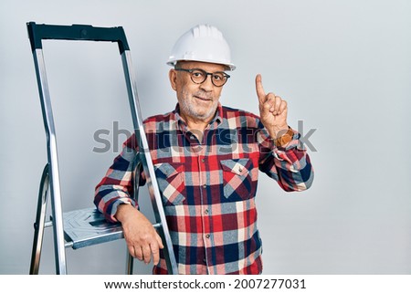 Handsome mature handyman close to construction stairs wearing hardhat with a big smile on face, pointing with hand finger to the side looking at the camera. 