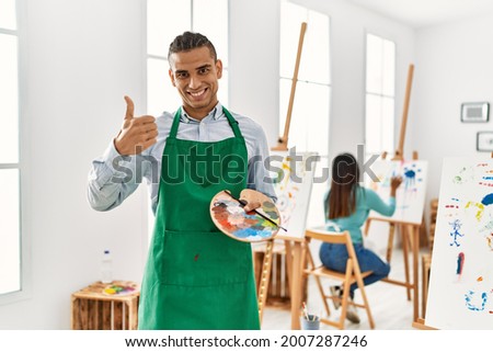 Young latin man standing at art studio smiling happy and positive, thumb up doing excellent and approval sign 